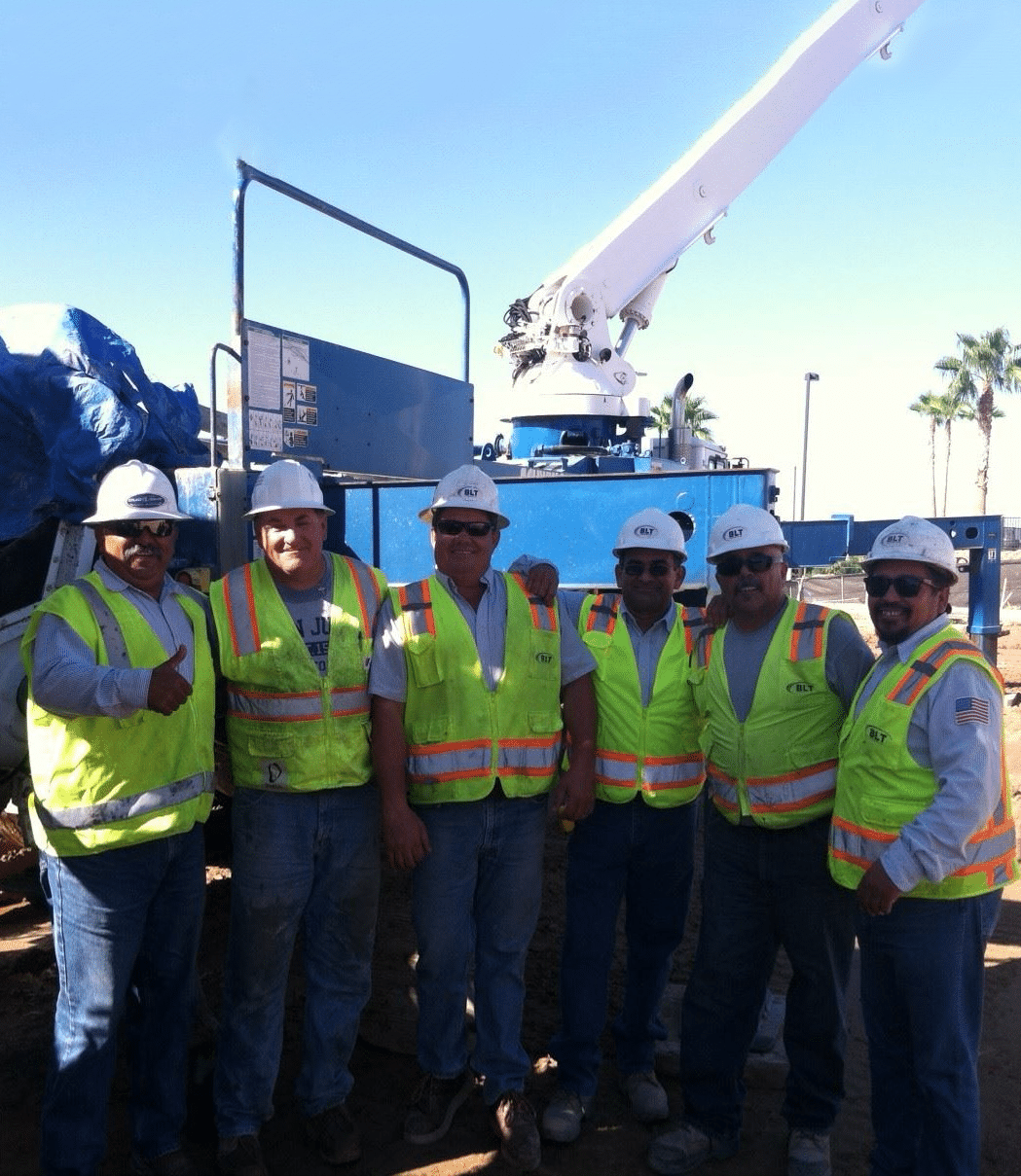 Five construction workers in safety gear and hard hats stand in front of equipment at a construction site on a clear day. Five construction workers in safety gear and hard hats stand in front of equipment at a construction site on a clear day.