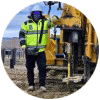 A person in a high-visibility jacket operates construction equipment at a work site with metal posts in the background.