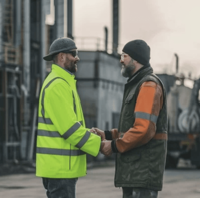 Two men in safety gear stand facing each other outside an industrial setting. One wears a bright yellow jacket, the other an orange and green jacket. They hold hands and converse. Two men in safety gear stand facing each other outside an industrial setting. One wears a bright yellow jacket, the other an orange and green jacket. They hold hands and converse.