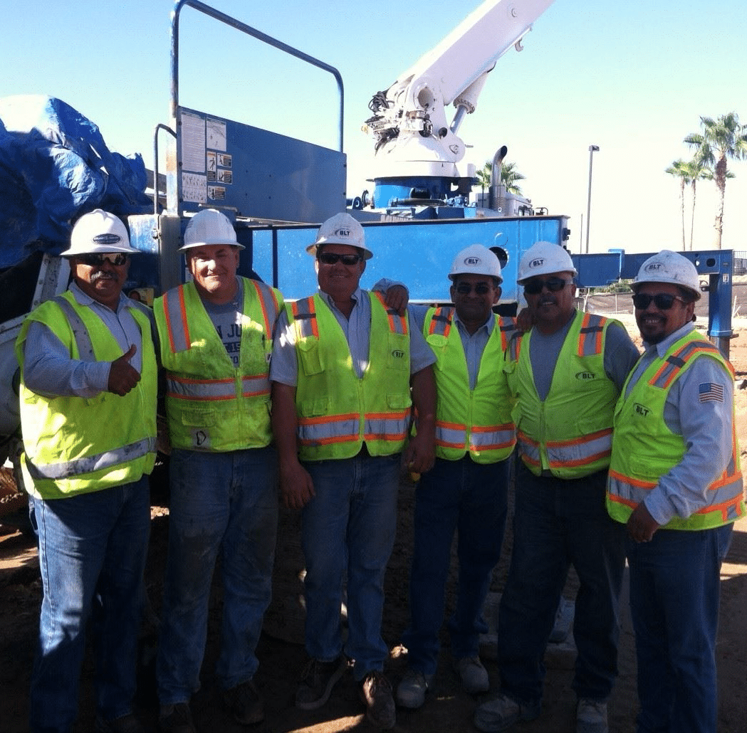 Six construction workers in safety vests and hard hats stand side by side at a work site with machinery in the background. Six construction workers in safety vests and hard hats stand side by side at a work site with machinery in the background.
