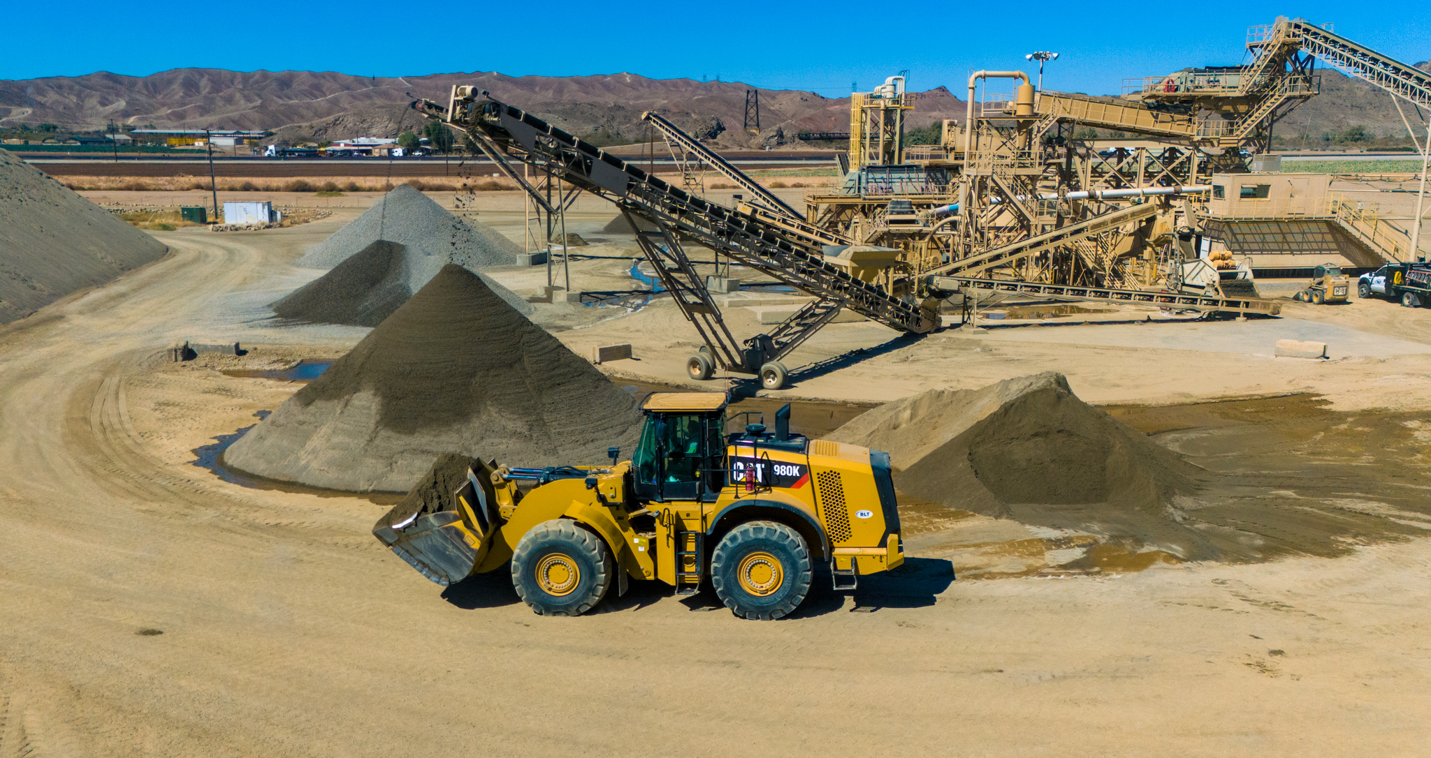A yellow bulldozer moves sand in a quarry with conveyor belts and piles of gravel under a clear blue sky. A yellow bulldozer moves sand in a quarry with conveyor belts and piles of gravel under a clear blue sky.